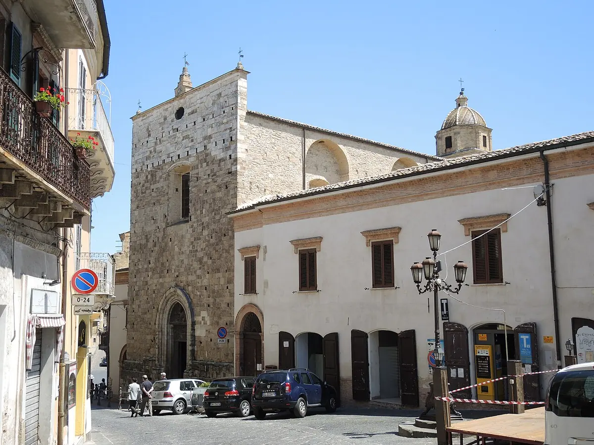 The tall, plain stone facade and bell tower of a medieval church beside a white building with green shutters, on a sunny town street with streetlamps
