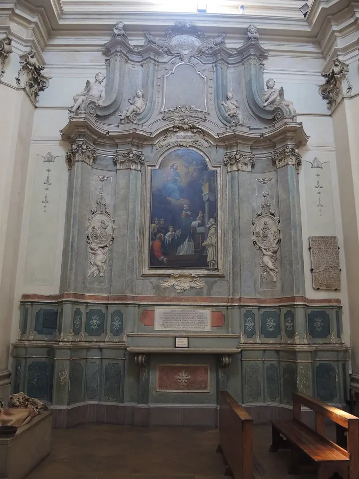 A tall pale blue and gray Baroque side altar with carved stucco frames around a painting, a marble inscription panel below and wooden pews in front