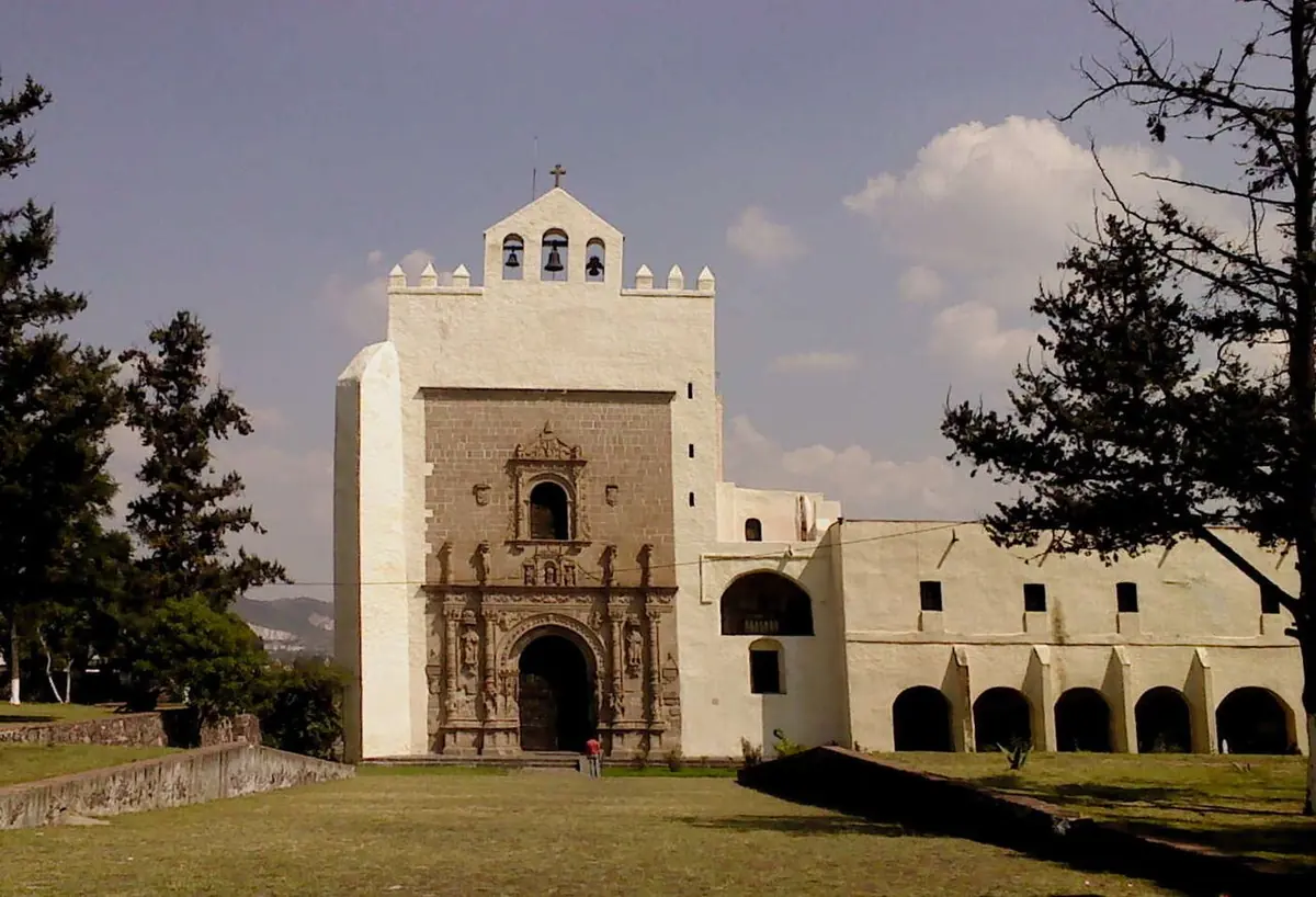 Whitewashed 16th-century church with a carved stone Plateresque doorway and a bell gable, beside a two-story arcaded convent wing, seen across a lawn
