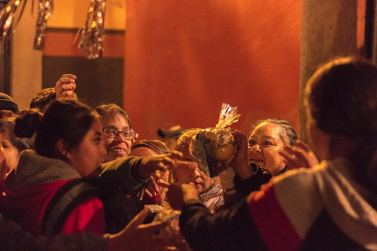 Smiling people in a crowded street at night reaching for small cellophane bags of candy and fruit handed out from a doorway