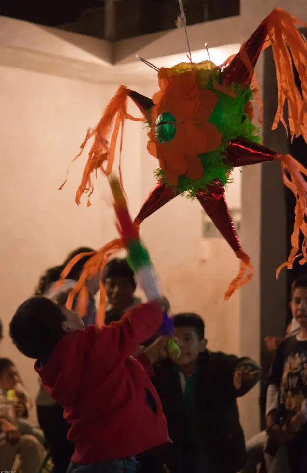 A boy in a red jacket swings a stick at a bright orange and green star piñata with paper streamers hanging from its cone-shaped points, while other children watch