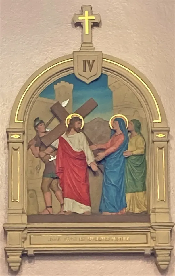 A carved and painted Station of the Cross on a church pillar showing Jesus carrying his cross and meeting his mother, with a small cross and number above