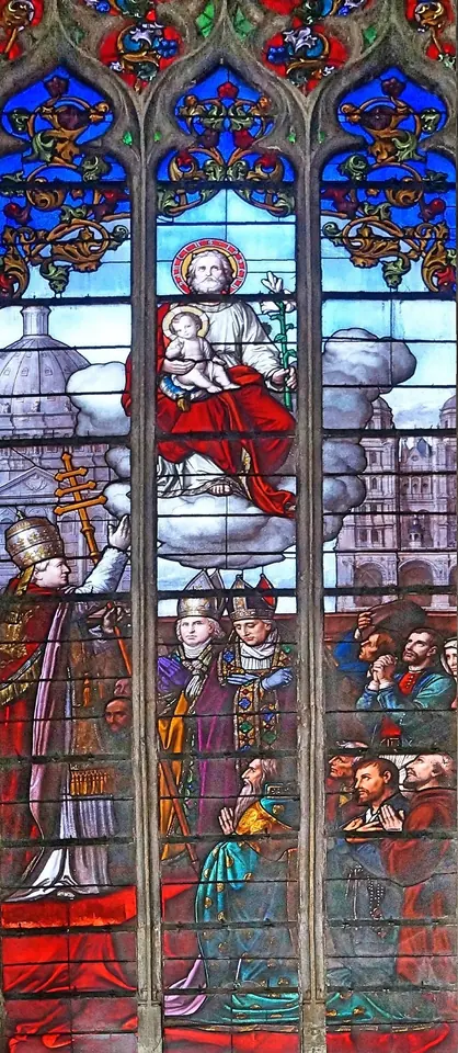 Tall stained-glass window of St. Joseph enthroned on clouds holding the Child Jesus, above a pope, bishops and faithful gathered in prayer