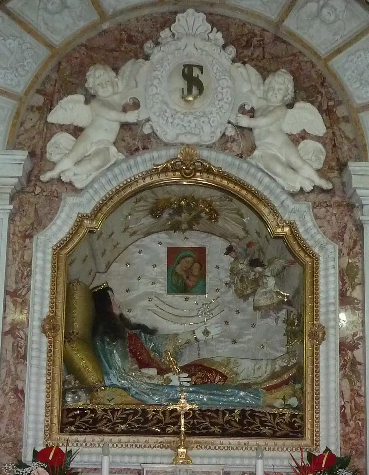 A reclining figure of a young girl in a pale blue and pink gown inside an ornate glass shrine framed by white stucco angels