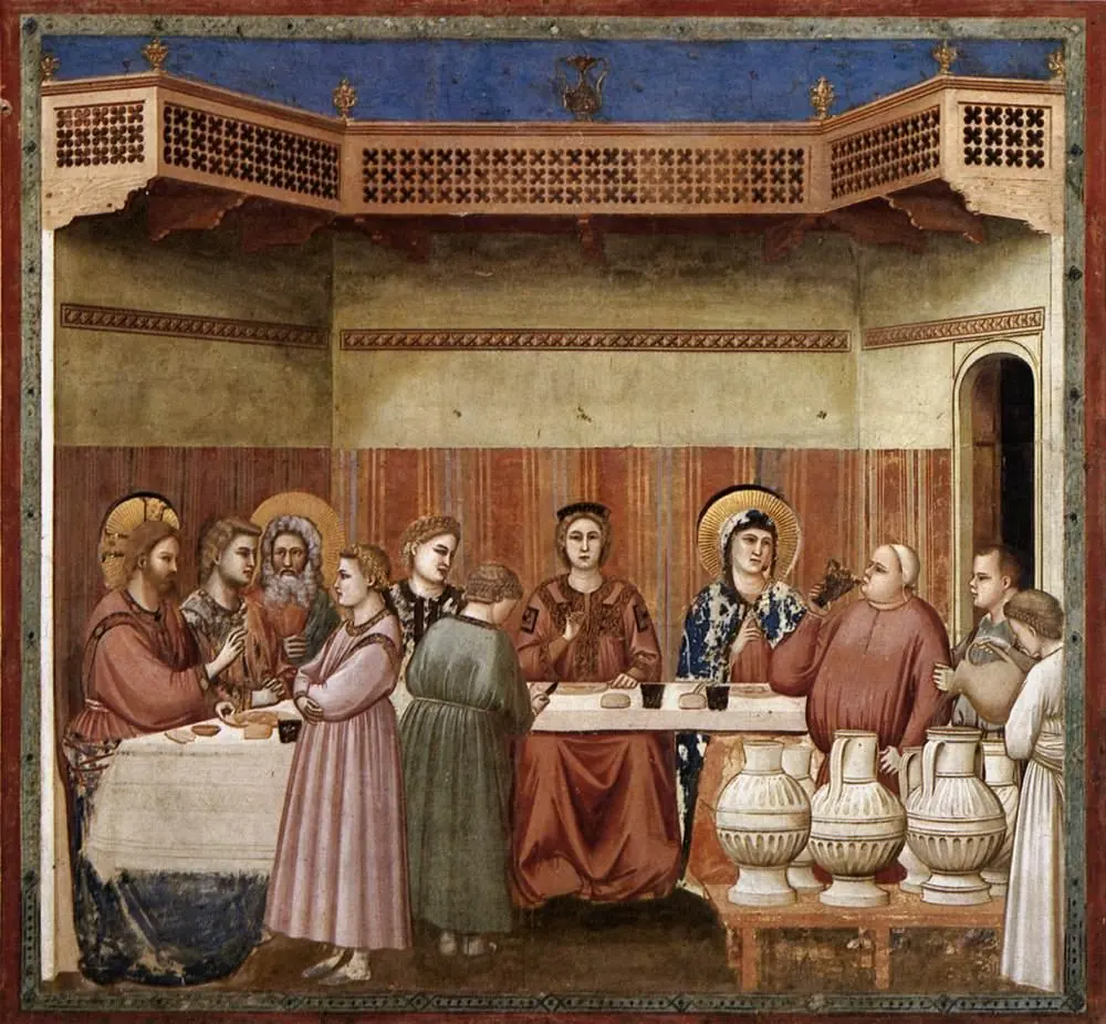 Wedding guests sit at a long table under a carved wooden balcony while Jesus raises his hand in blessing and a servant tastes wine beside a row of large stone jars