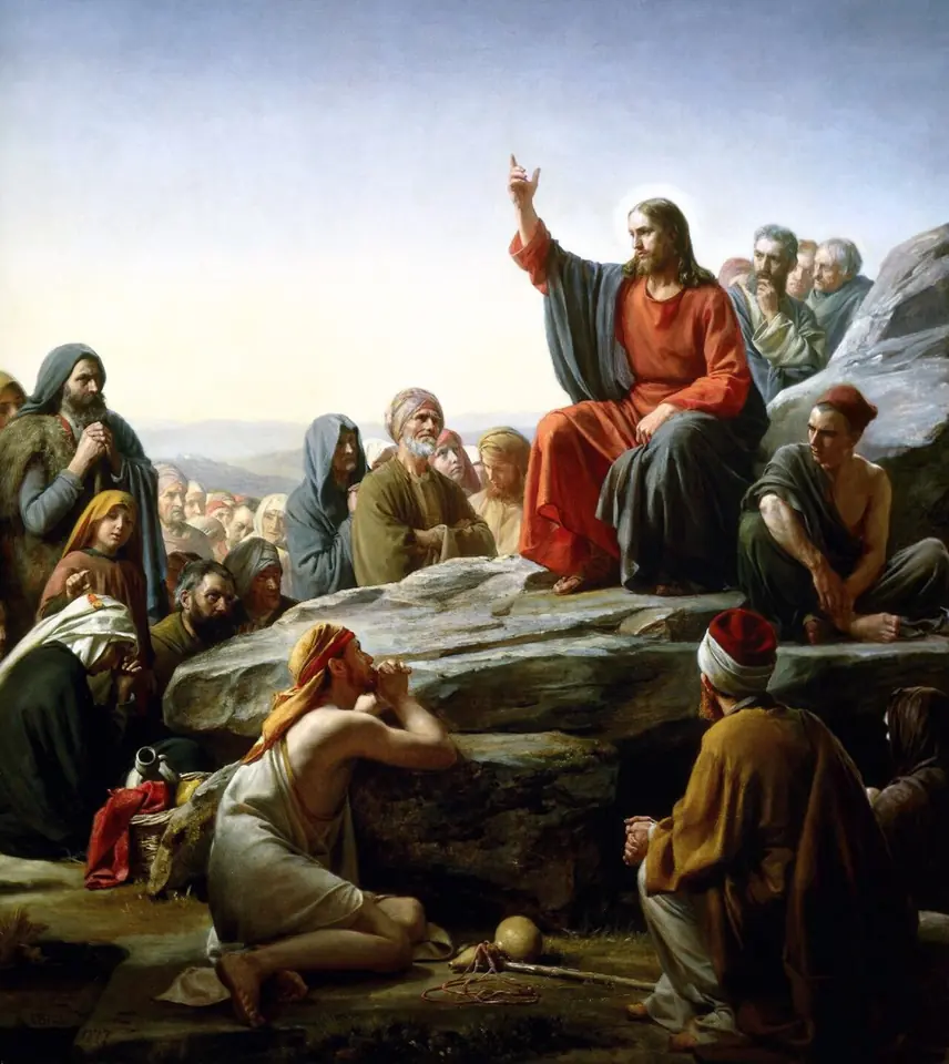 Jesus in a red robe sits on a rock on a hillside, one hand raised as he teaches a crowd of men, women and children gathered on the slope around him
