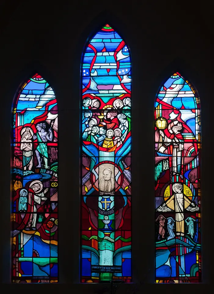 A tall three-light stained-glass window in deep blues and reds showing small scenes from the life of Christ, with a portrait of Pope John Paul II in the center