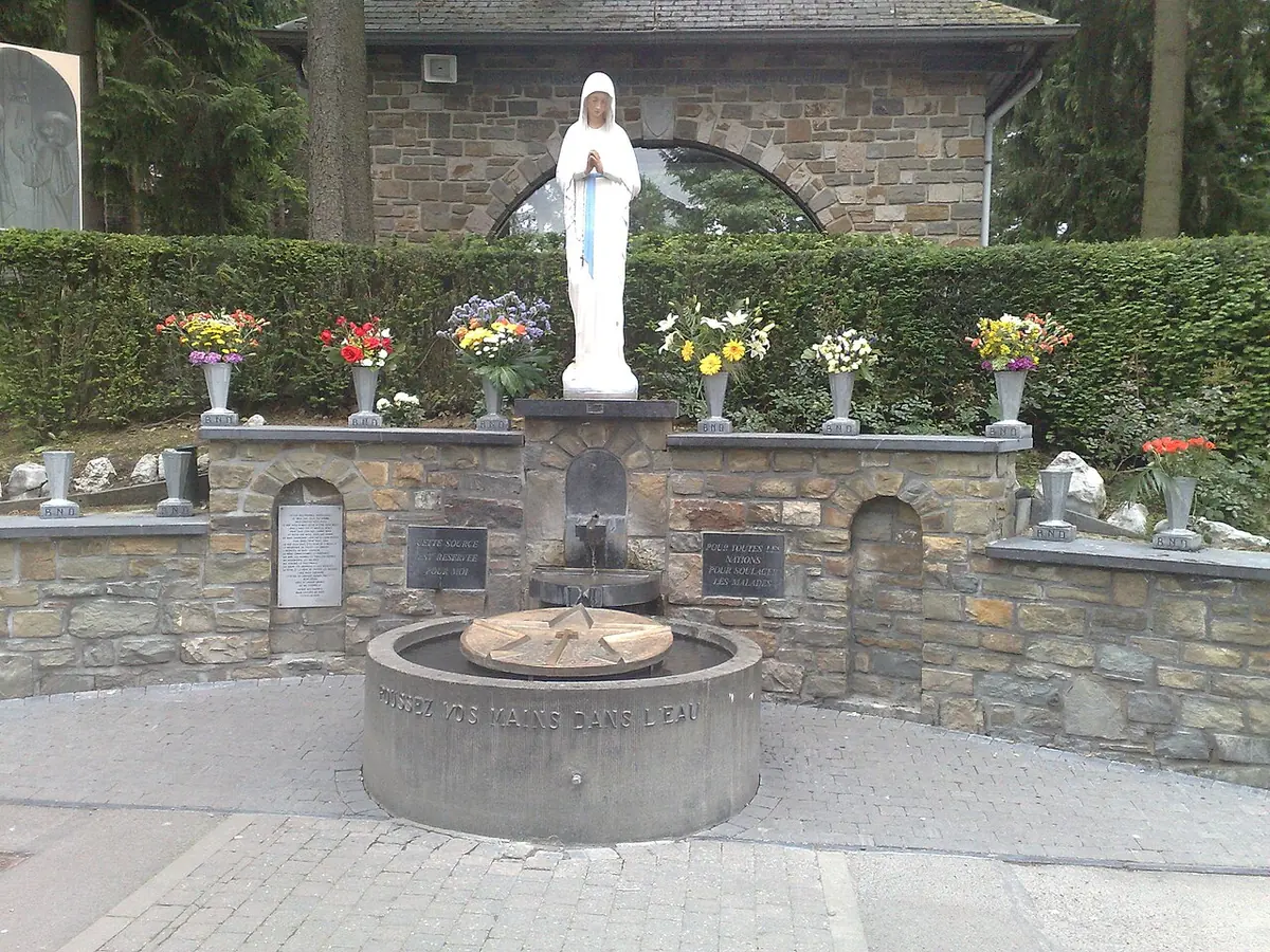 A white statue of the Virgin above a low stone wall with flowers and a round basin fed by a spring
