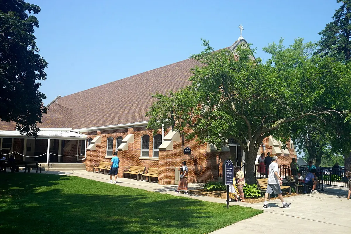 A red-brick chapel with a steep roof and a small white cross, with pilgrims walking along a path under a large shade tree
