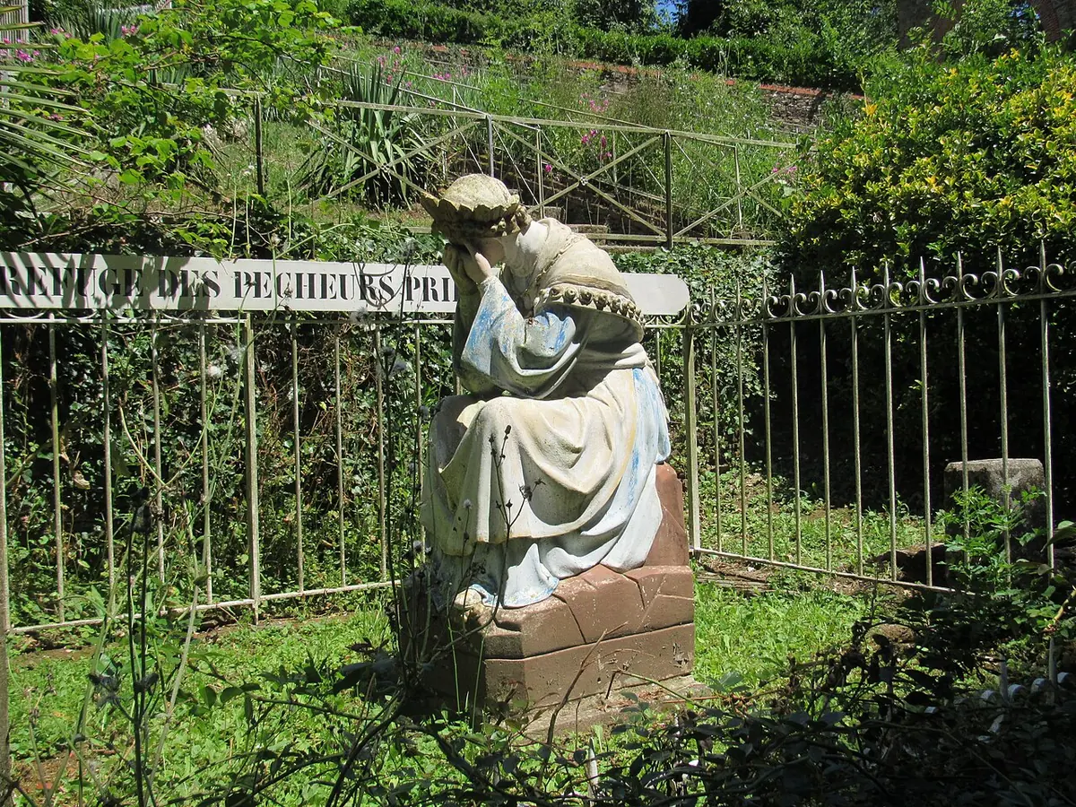 White statue of the Virgin seated on a stone with her face in her hands, weeping, in a fenced garden on the mountain
