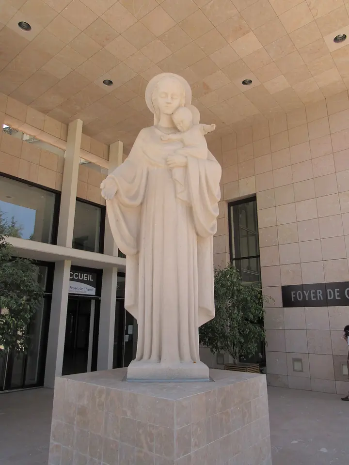 White stone statue of the Virgin Mary holding the Christ Child on her arm, standing on a pedestal in a bright modern entrance hall