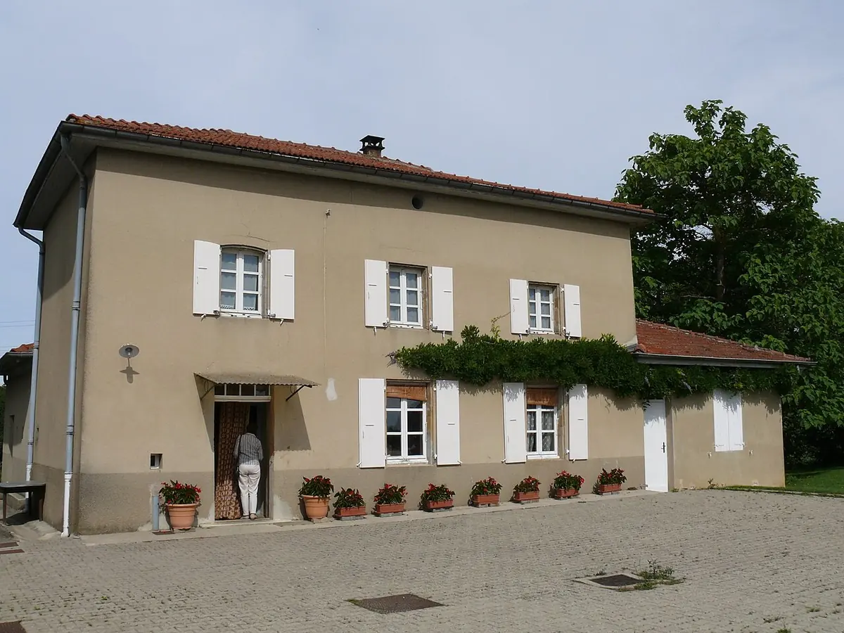 Plain two-story farmhouse with white shutters and a red tile roof, pots of red flowers along the front wall and a visitor at the open door