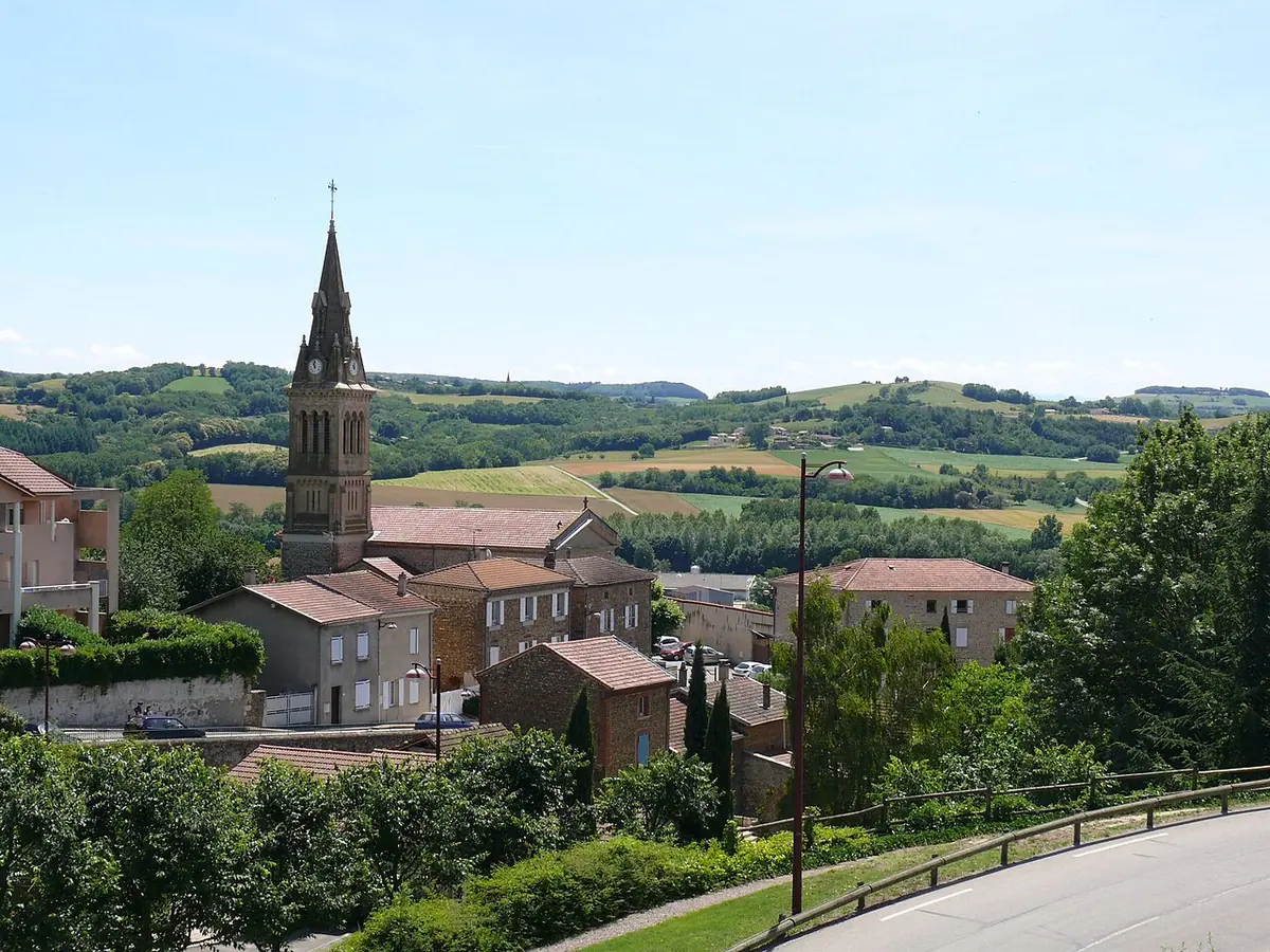 Village church with a tall pointed stone bell tower above red tile roofs, with green hills and fields stretching behind