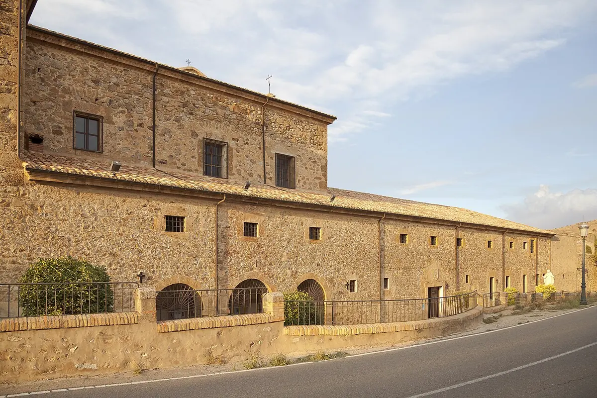 The long stone wall of a Spanish convent with small barred windows and arched doors along a road under a pale evening sky