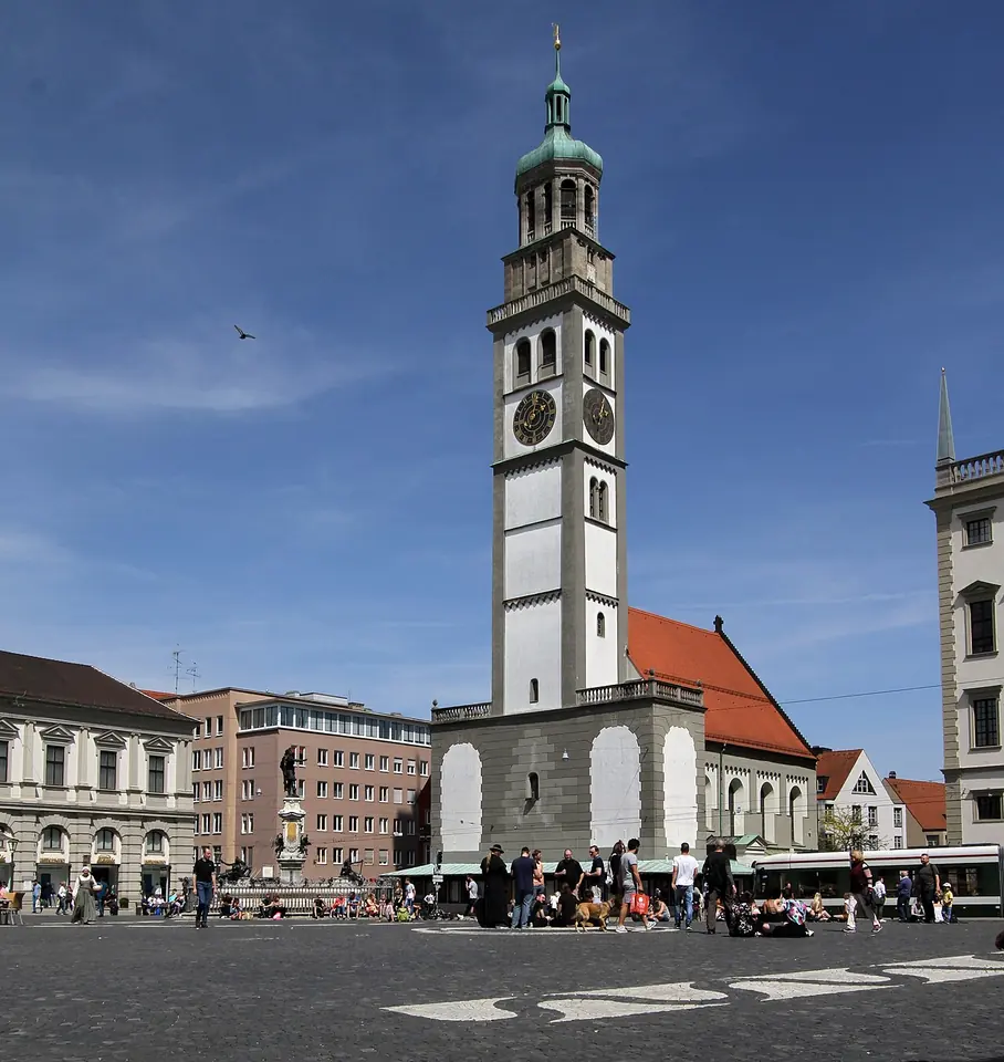 Alta torre blanca con cúpula verde en forma de cebolla junto a una pequeña iglesia de tejado rojo, en una plaza de Augsburgo bajo el cielo azul