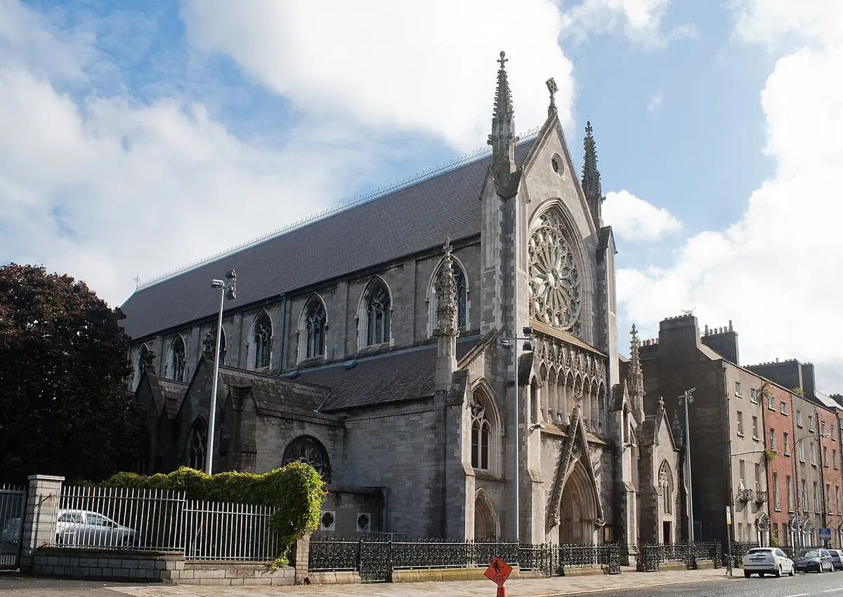 Gray stone Gothic Revival church with a rose window and pointed spires, beside a row of brick Georgian houses on a Dublin street