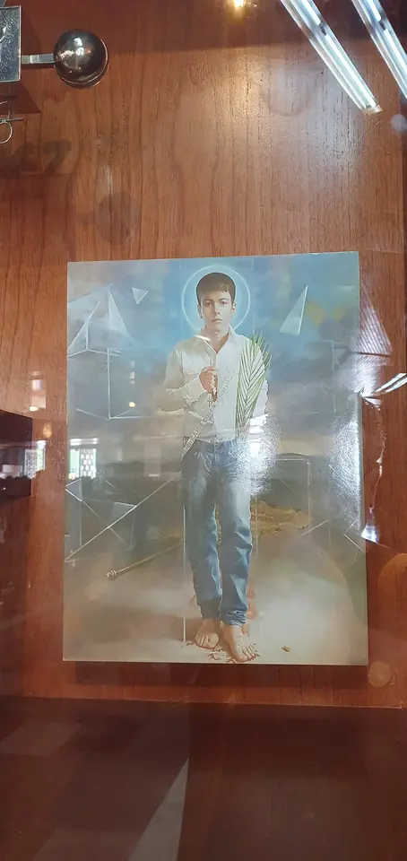 Painted image of a teenage boy in a white shirt and jeans with a halo, holding a cross, displayed behind glass in a wooden shrine