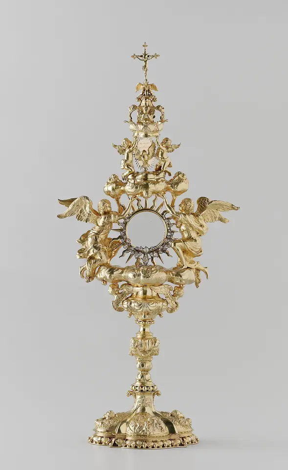 A gilded silver Baroque monstrance with a round window framed by short rays, two kneeling angels with spread wings, scrolling leaves, a cross on top and a lobed foot
