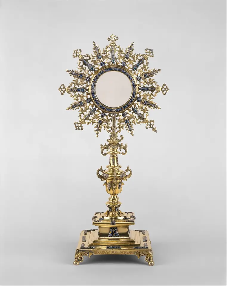 A silver-gilt sunburst monstrance with pointed and flame-shaped rays set with dark enamel, a round glass window at the center, a vase-shaped stem and a square footed base