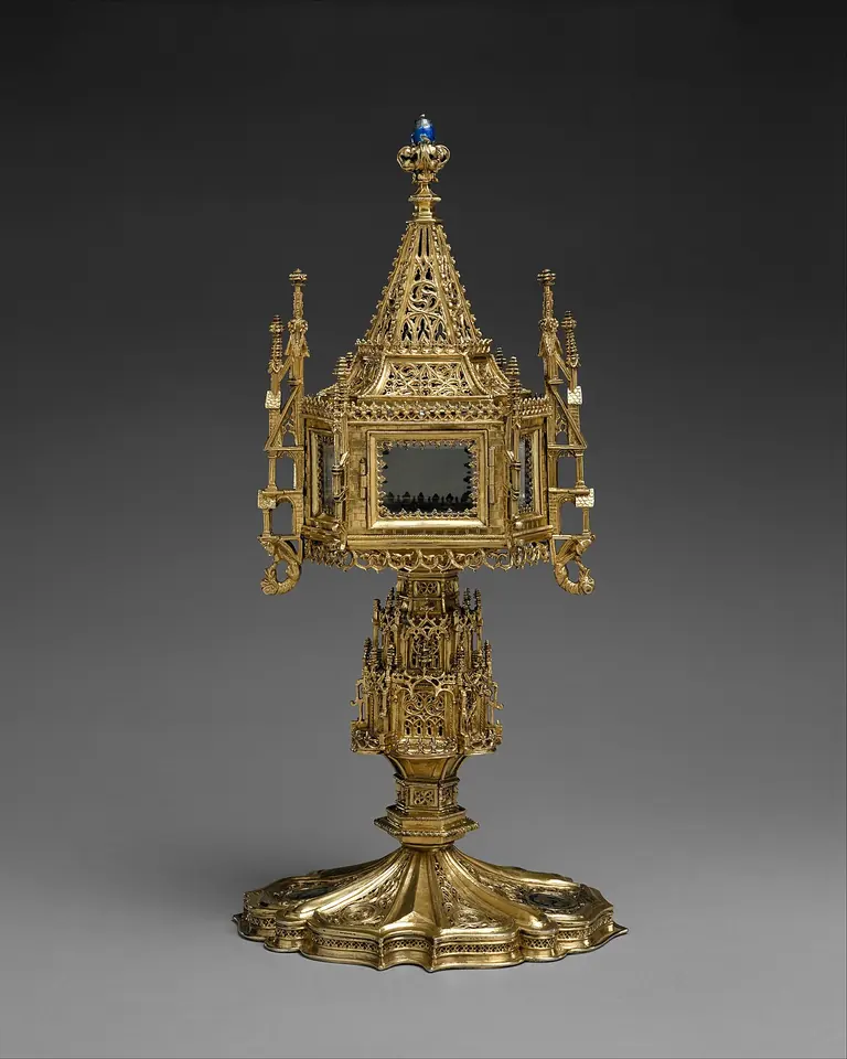 A Gothic gilded silver monstrance shaped like a little tower, with pinnacles and a spire around a glass window, on a stem with a lobed base, against a dark background