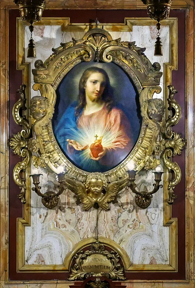 Oval painting of Christ in a red robe and blue mantle holding a glowing heart crowned with a small cross, set in an ornate gilded frame on a marble wall