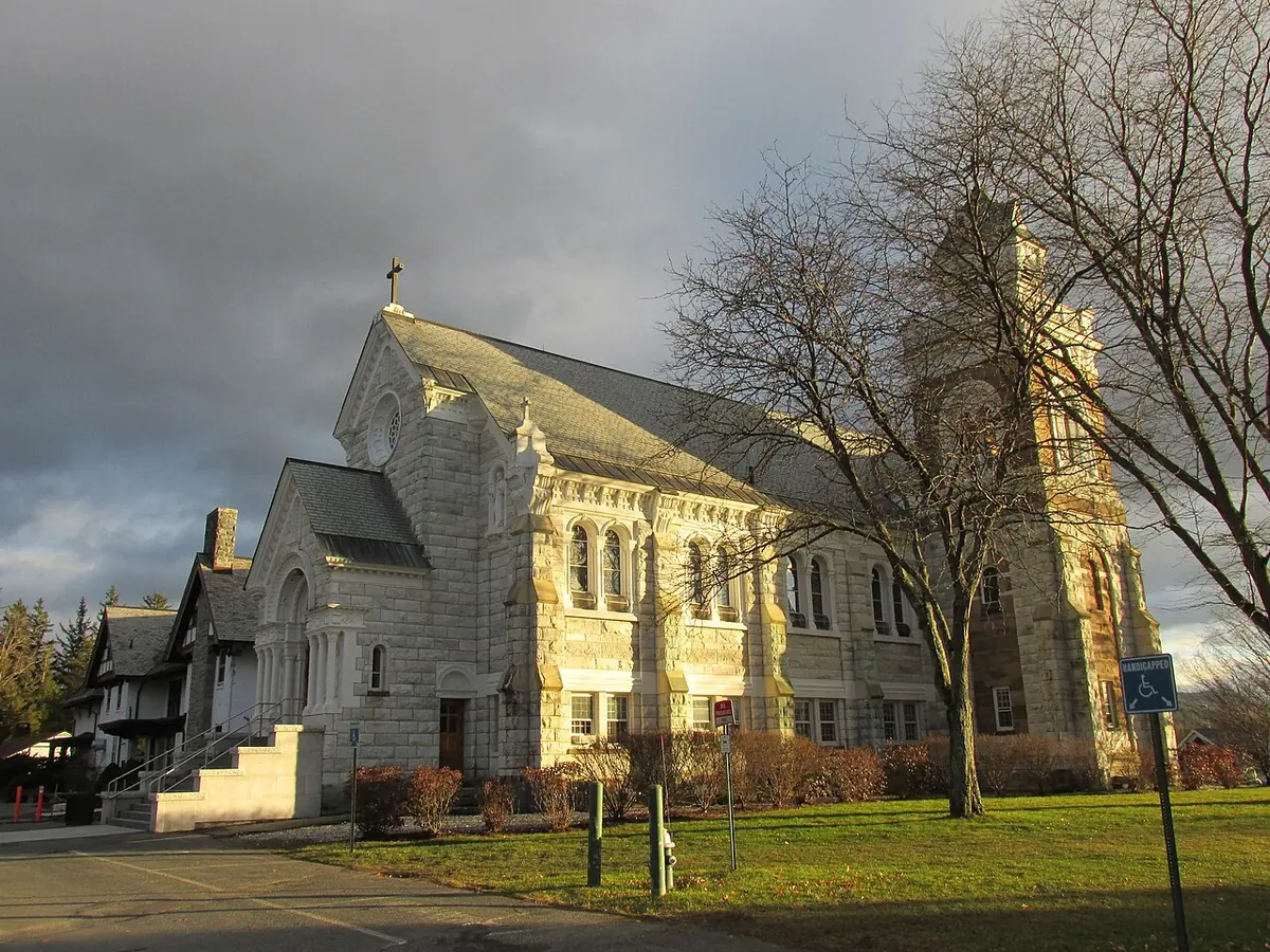 A pale stone church with steep roofs and a square tower, seen from the side across a lawn under a stormy sky, with bare trees around it