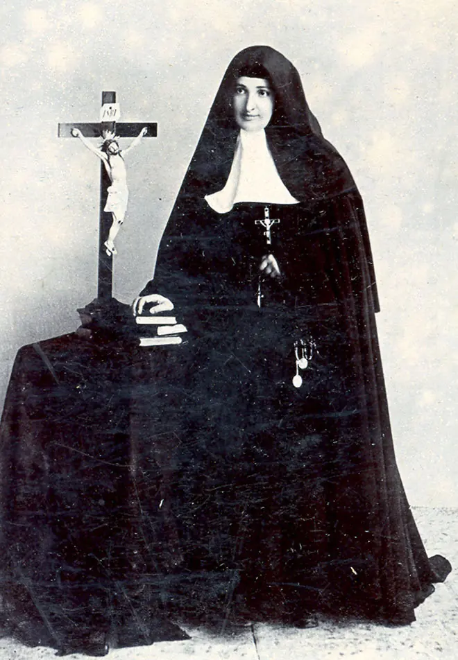 Antigua fotografía en blanco y negro de una monja con hábito negro y toca blanca, sentada junto a un crucifijo alto