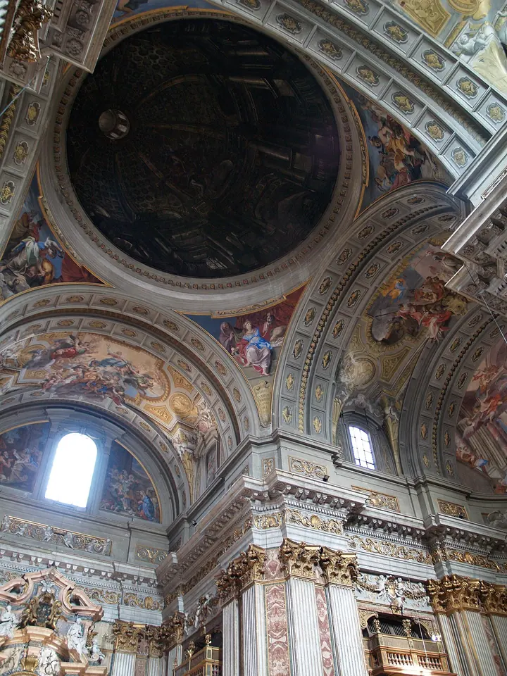 Vista hacia lo alto del interior barroco de una iglesia, con columnas de mármol, arcos con frescos y una cúpula pintada