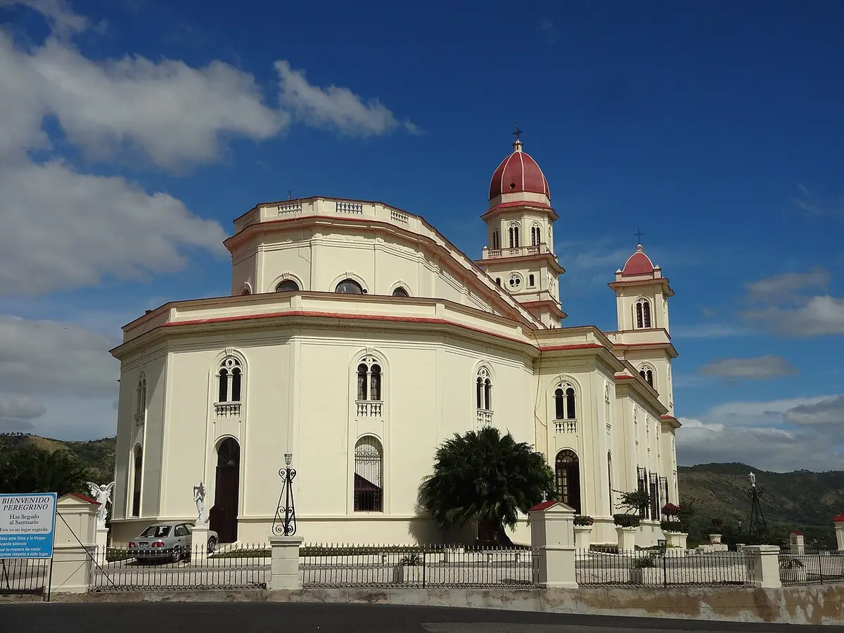 Cream-colored basilica with a central bell tower and two smaller towers with red domes, standing on a hill under a blue sky
