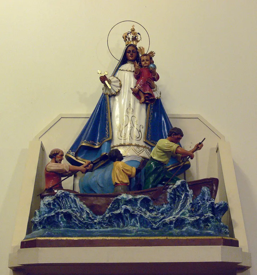 Painted sculpture of the crowned Virgin in white and blue holding the Child above three men rowing a small boat through blue waves