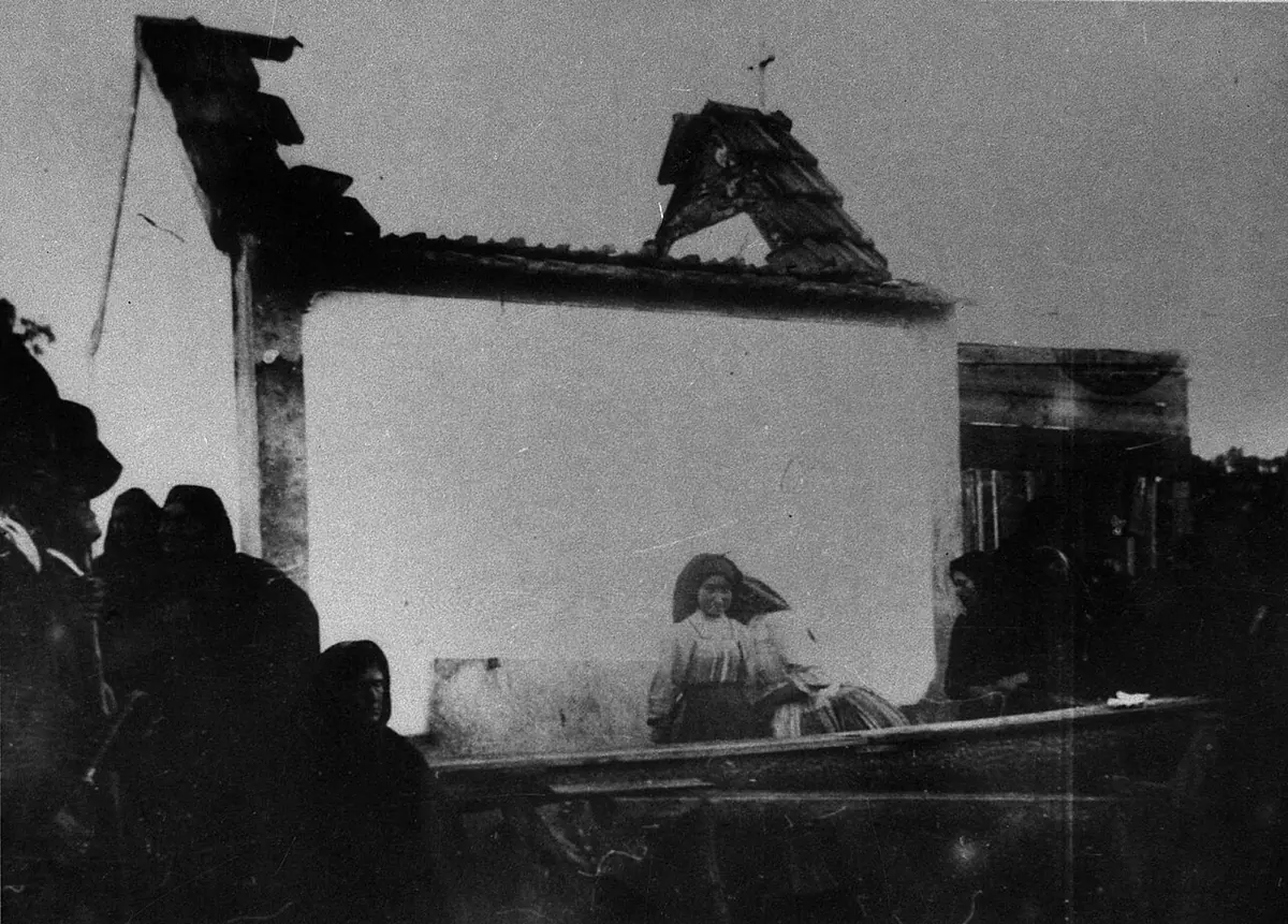 Old photograph of the little chapel with its roof blown off, as women in dark shawls sit and kneel in front of it
