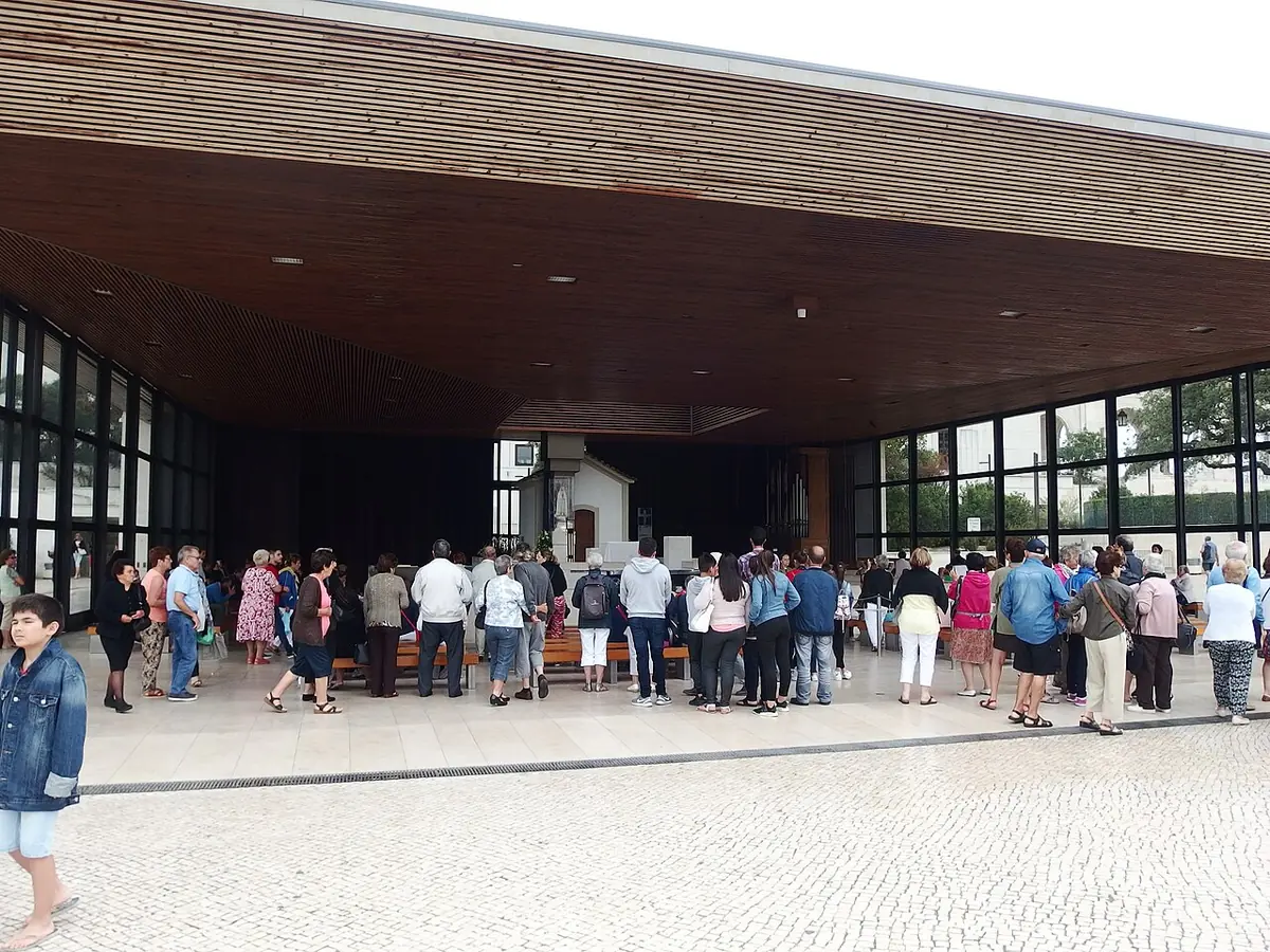 Pilgrims standing under a modern open porch with a wooden ceiling and glass walls that shelters the small white Chapel of the Apparitions