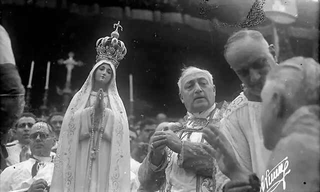 Photograph of a cardinal in vestments beside the statue of Our Lady of Fátima wearing a tall jeweled crown, surrounded by clergy