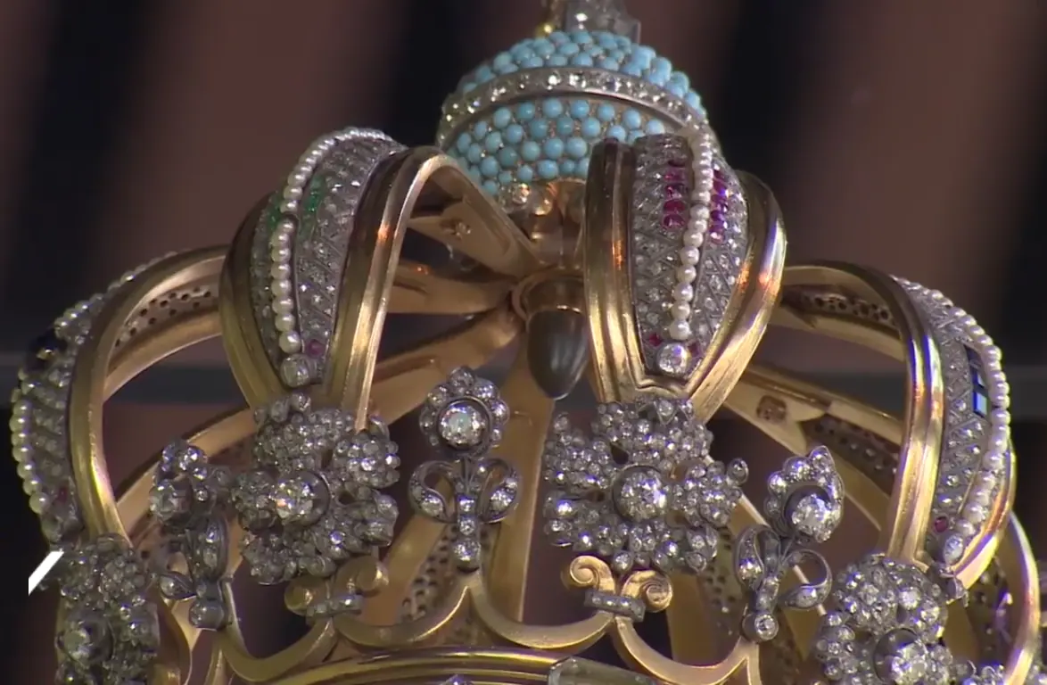 Close view of a gold crown set with pearls and diamonds, with a small dark bullet fixed where the arches meet beneath a turquoise globe