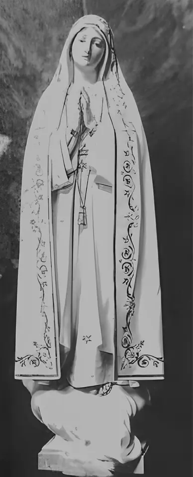 Old photograph of a carved wooden statue of the Virgin in a long white mantle with ornamented borders, hands joined with a rosary, standing on a cloud