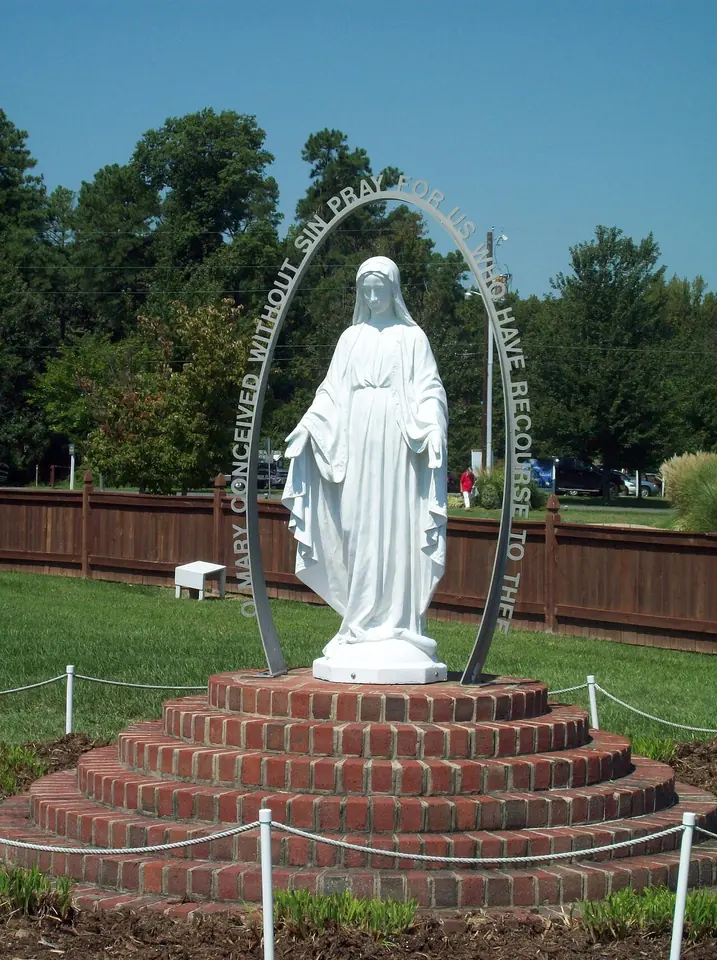 White outdoor statue of Mary with arms lowered and hands open, standing on a globe inside a large oval ring bearing the words "O Mary conceived without sin, pray for us who have recourse to thee," on a round brick base