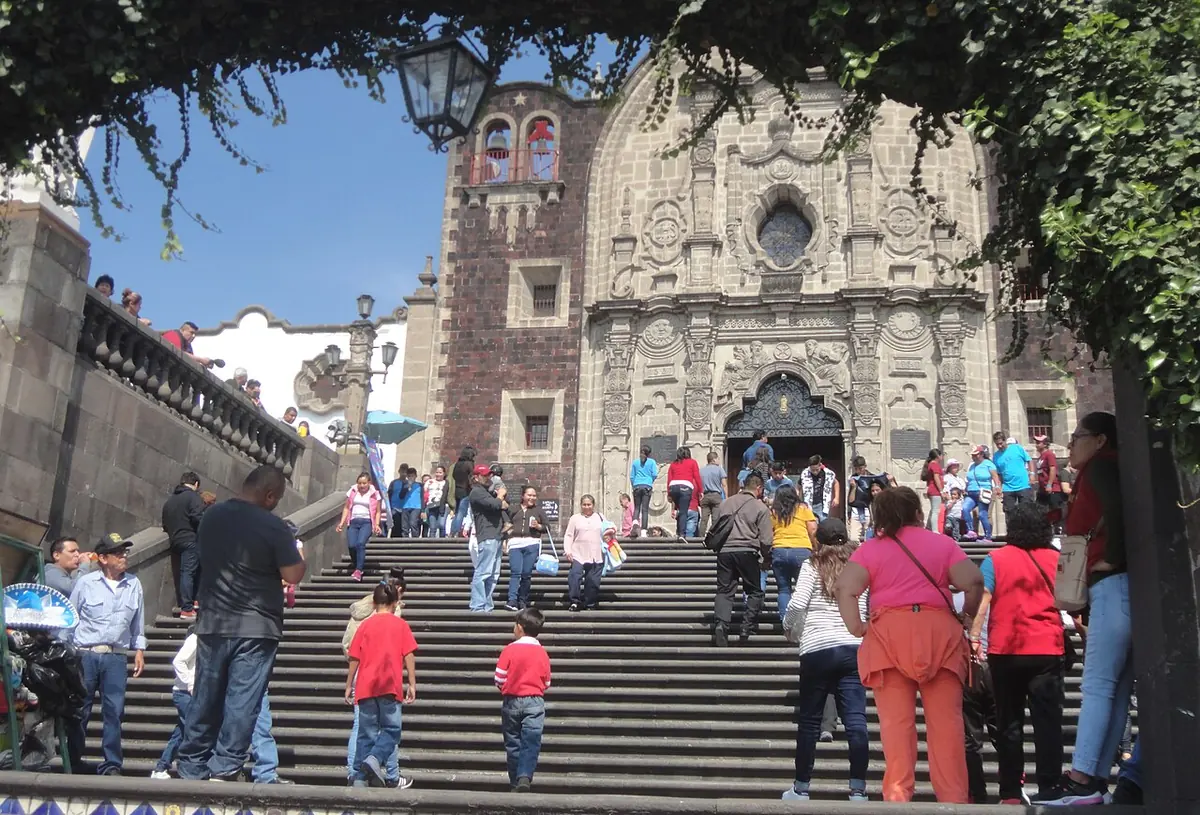 Visitantes que suben una amplia escalinata de piedra hacia la fachada labrada de la capilla en la cumbre del cerro del Tepeyac