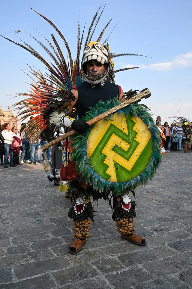 Danzante con un alto penacho de plumas y traje de inspiración azteca, con un escudo redondo de plumas verdes, en la explanada de la basílica