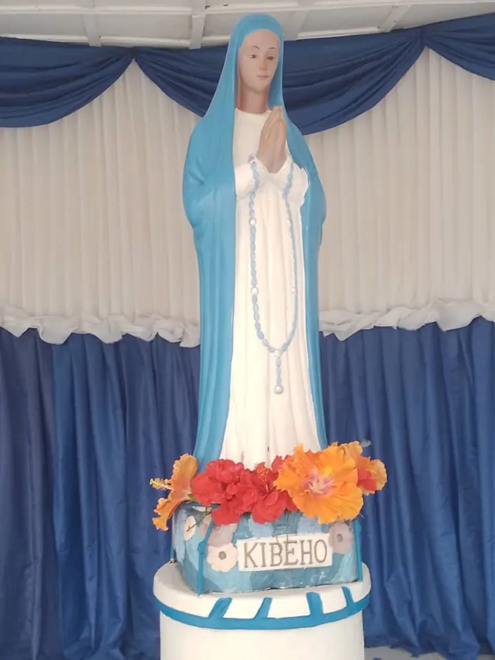A statue of Mary in a white robe and long blue veil, hands joined in prayer with a white rosary, standing on a pedestal decorated with red and orange flowers and the word Kibeho