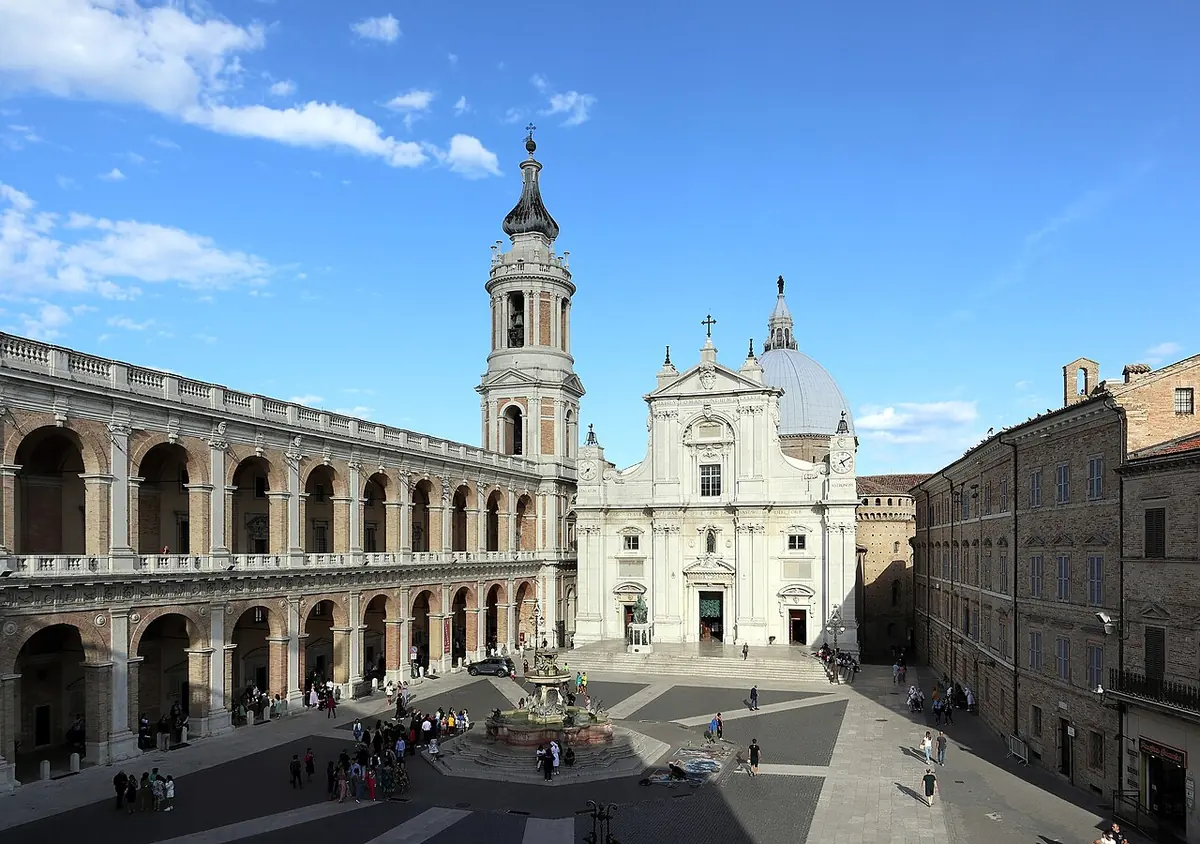 Sunny view across the Piazza della Madonna in Loreto to the white facade, bell tower and dome of the Basilica of the Holy House, with the arcaded Apostolic Palace on the left