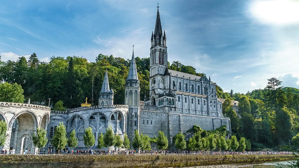 Basílica gótica de color claro con una alta aguja sobre laderas arboladas, con la basílica del Rosario, de cúpula, y sus rampas curvas a sus pies junto a un río