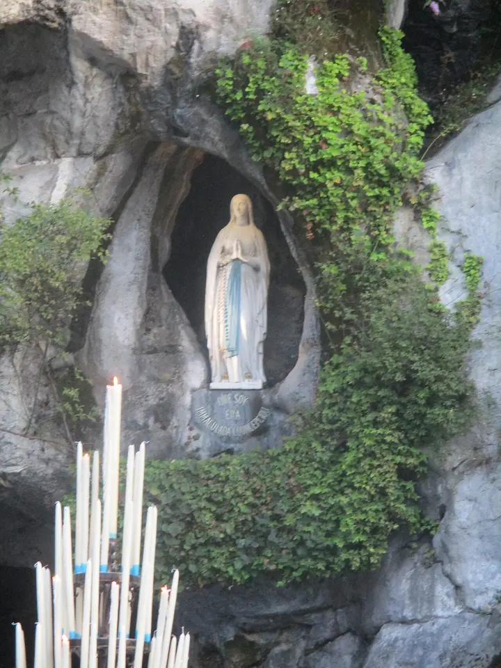Estatua blanca con cinta azul en un nicho ovalado de roca gris cubierta de hiedra y helechos, con altos cirios blancos encendidos abajo