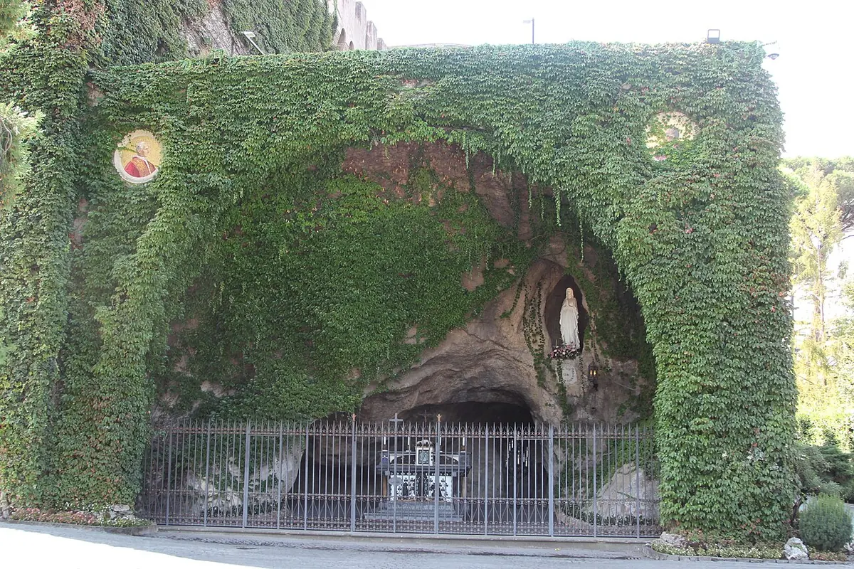 Gran gruta artificial de roca cubierta de hiedra verde, con una estatua blanca de la Virgen en un nicho y un altar enrejado en el interior de la cueva