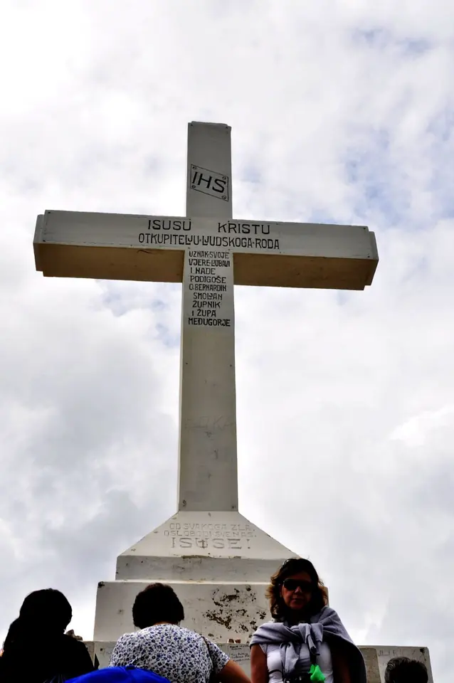 Una gran cruz blanca de hormigón con una inscripción en croata a Jesucristo Redentor, vista desde abajo, con peregrinos que descansan a su pie