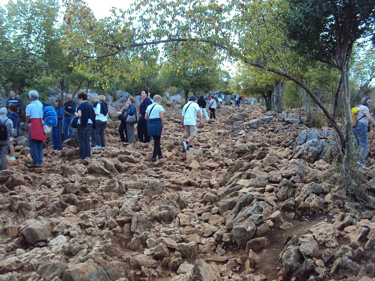 Peregrinos que suben por un sendero empinado de rocas rojizas y afiladas entre pequeños árboles