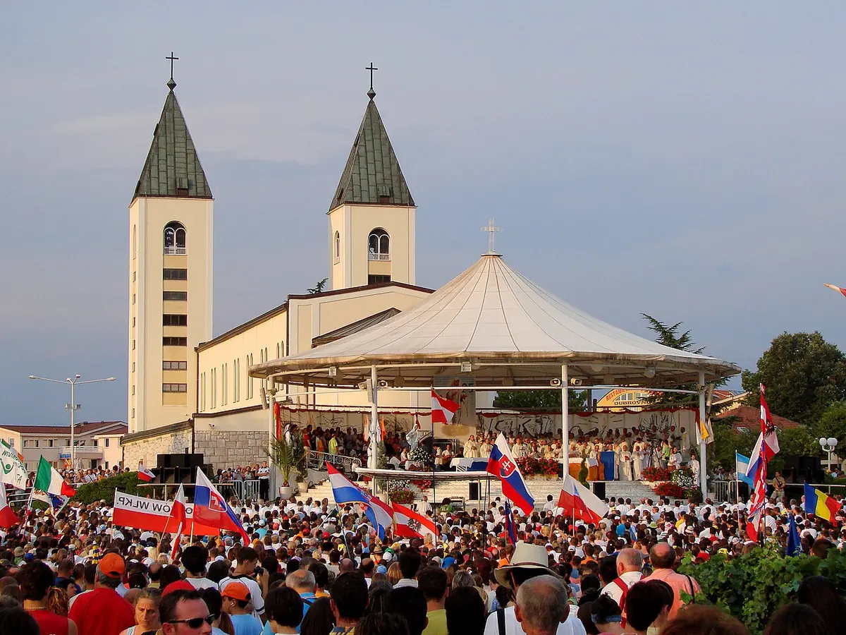 Una enorme multitud de jóvenes que agitan banderas de muchas naciones frente a una iglesia de dos torres y un toldo blanco sobre el altar
