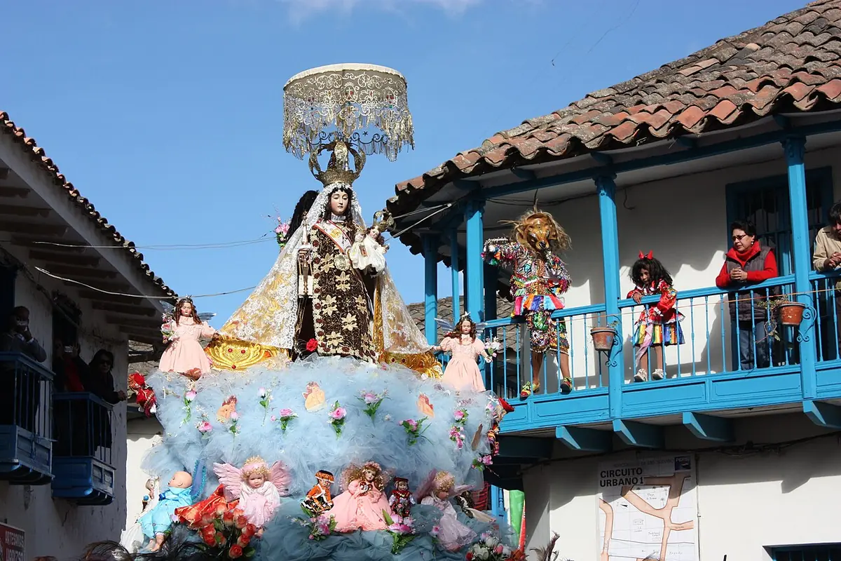 A richly dressed statue of the Virgen del Carmen under a silver canopy, on a float covered with pale blue cloth and dolls of angels, passing a house with a blue balcony in Paucartambo