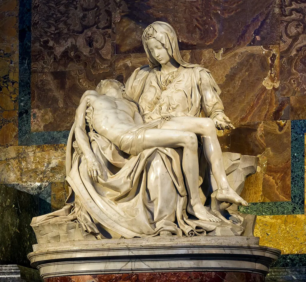 White marble sculpture of a young Virgin Mary seated with the body of the dead Christ lying across her lap, her left hand open, in a marble niche
