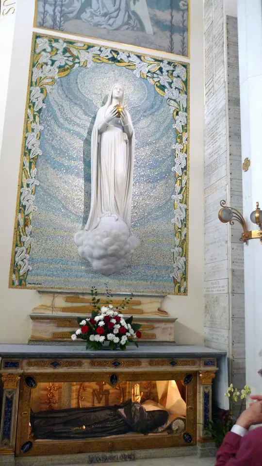 A white statue of the Virgin holding a globe against a blue mosaic, above an altar with a glass case in which the body of a nun in a dark habit lies