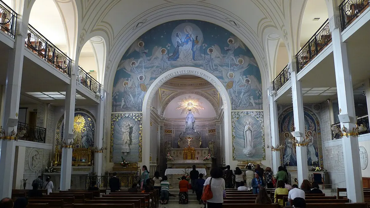Interior of a white chapel with balconies on both sides, pews full of visitors and a blue and gold mosaic apse around a white statue of the Virgin above the high altar